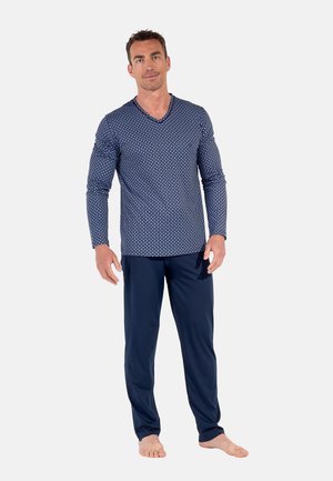 Ein Mann steht barfuß und trägt ein marineblaues, gemustertes Langarmshirt mit V-Ausschnitt und eine unifarbene marineblaue Hose vor einfachem Hintergrund.