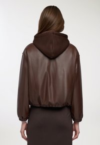 Giacca cropped in finta pelle marrone con cappuccio marrone scuro, polsini e vita elasticizzati, caratterizzata da una texture liscia e da un design minimale.