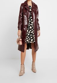 Vinröd glansig trenchcoat med breda revers, svart klänning med krämfärgad blommönster och beige ankelboots med kraftig klack.