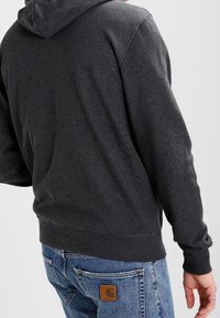 Sweat-shirt à capuche gris charbon avec un tissu texturé, des poignets et une taille élastiques. Détail avec un petit écusson logo au dos.