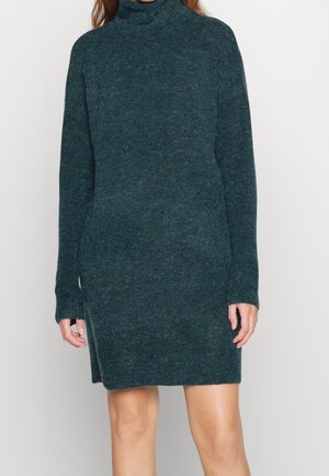 Femme portant une robe-pull à col roulé vert foncé à manches longues, debout devant un fond clair uni.