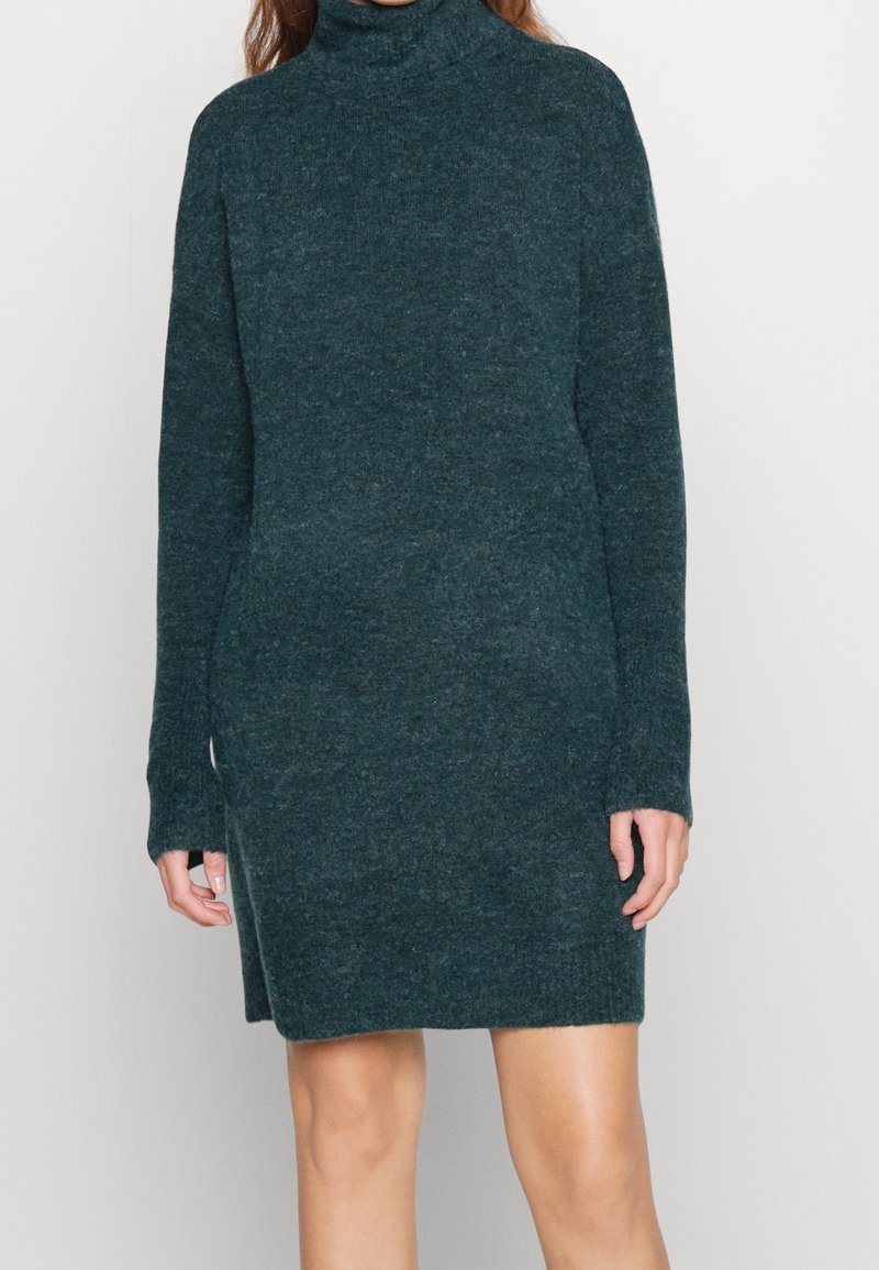 Femme portant une robe-pull à col roulé vert foncé à manches longues, debout devant un fond clair uni.