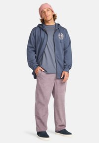 Felpa chiusa di colore blu scuro con logo, abbinata a una t-shirt grigia e pantaloni in velluto a coste leggeri di colore viola chiaro, completata da scarpe blu navy e un berretto rosa.