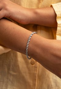 Bracciale a catena a corda intrecciata in argento indossato su un polso con una camicia button-up marrone chiaro sullo sfondo.