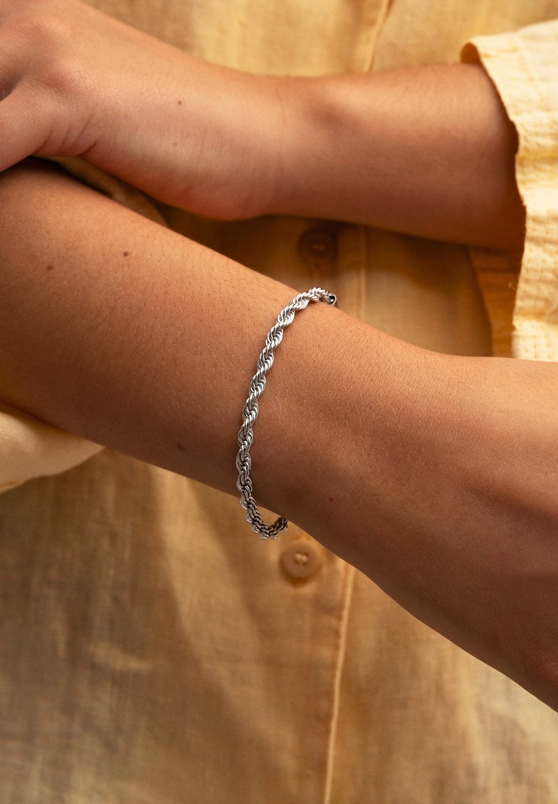 Bracciale a catena a corda intrecciata in argento indossato su un polso con una camicia button-up marrone chiaro sullo sfondo.