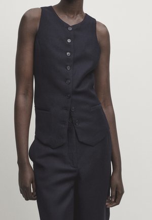 Gros plan d'une personne portant un gilet noir sans manches boutonné et un pantalon noir assorti sur un fond uni.