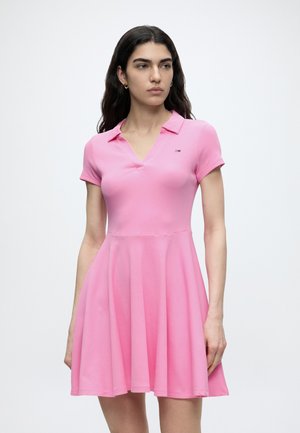 Vrouw draagt een roze jurk met korte mouwen en kraag, een aansluitend lijfje en een uitlopende rok, staand tegen een effen witte achtergrond.