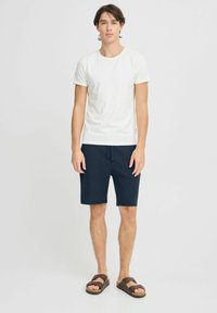Fehér pamut póló, navy kék struktúrált rövidnadrággal, rugalmas derékkal, és barna szandálokkal, állítható pántokkal.
