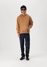 Sudadera marrón con un bolsillo tipo canguro, combinada con jeans azul oscuro. El modelo lleva gafas de sol negras y zapatillas negras, de pie frente a un fondo liso.