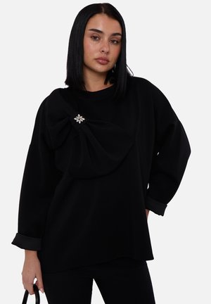 Black sweatshirt with loose fit, featuring a large bow detail on the front adorned with a floral brooch. Long sleeves with rolled cuffs.
