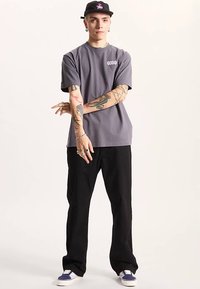 Grå kortärmad t-shirt med logotyp, kombinerad med svarta vida byxor och sneakers med lila detaljer, bärs med en svart keps.