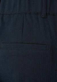 Tmavě modré kalhoty s herringbonovým vzorem, viditelnou zadní kapsou a upraveným šitím podél pasu.