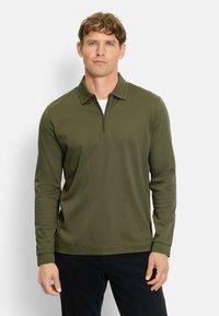 Dunkelgrünes Langarm-Polohemd mit einem Halb-Reißverschluss, das einen Kragen hat und aus einem glatten, strukturierten Stoff besteht. Stylisch kombiniert mit schwarzen Cordhosen.
