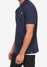 Muž v námořnickém modrém polo tričku s krátkými rukávy a malým bílým logem na hrudi a černých kalhotách.