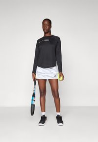 Zwarte lange mouwen sportshirt, witte gestreepte rok, zwarte tennisschoenen, met een tennisracket en bal in de hand, staand op een effen achtergrond.