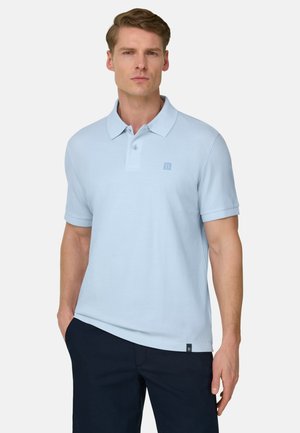 Mand iført lyseblå polo-shirt med korte ærmer og krave samt mørkeblå bukser, stående foran en ensfarvet hvid baggrund.