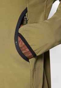 Maslinasto zelena jakna s elegantnim dizajnom, zipovima džepom s crnim obrubom i smešnim kožnim detaljem unutar džepa.