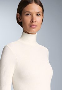 Crème col roulé trui gemaakt van textuur gebreide stof, met een getailleerd ontwerp en lange mouwen, tegen een neutrale achtergrond.