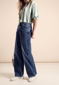 Blusa a rayas verdes y blancas, jeans de mezclilla azul de pierna ancha y zapatillas blancas. Textura informal y ajuste relajado. Bolsa de croché simple.