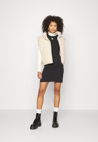Beige puffer vest with a high collar over a tight black dress. Black lace-up boots and white socks complete the look. Smooth textures, minimal design.