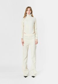 Conjunto color crema con una blusa de cuello alto y pantalones ajustados. Presenta detalles de costura y una textura suave y elástica; terminado con tacones altos negros en punta.