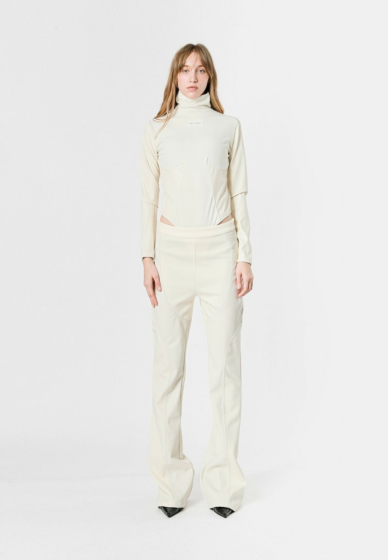 Conjunto color crema con una blusa de cuello alto y pantalones ajustados. Presenta detalles de costura y una textura suave y elástica; terminado con tacones altos negros en punta.