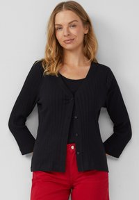 Zwarte geribde cardigan met een v-hals, lange mouwen en knopen aan de voorkant, gecombineerd met een zwarte top en rode broek. Soepele textuur en aansluitend ontwerp.