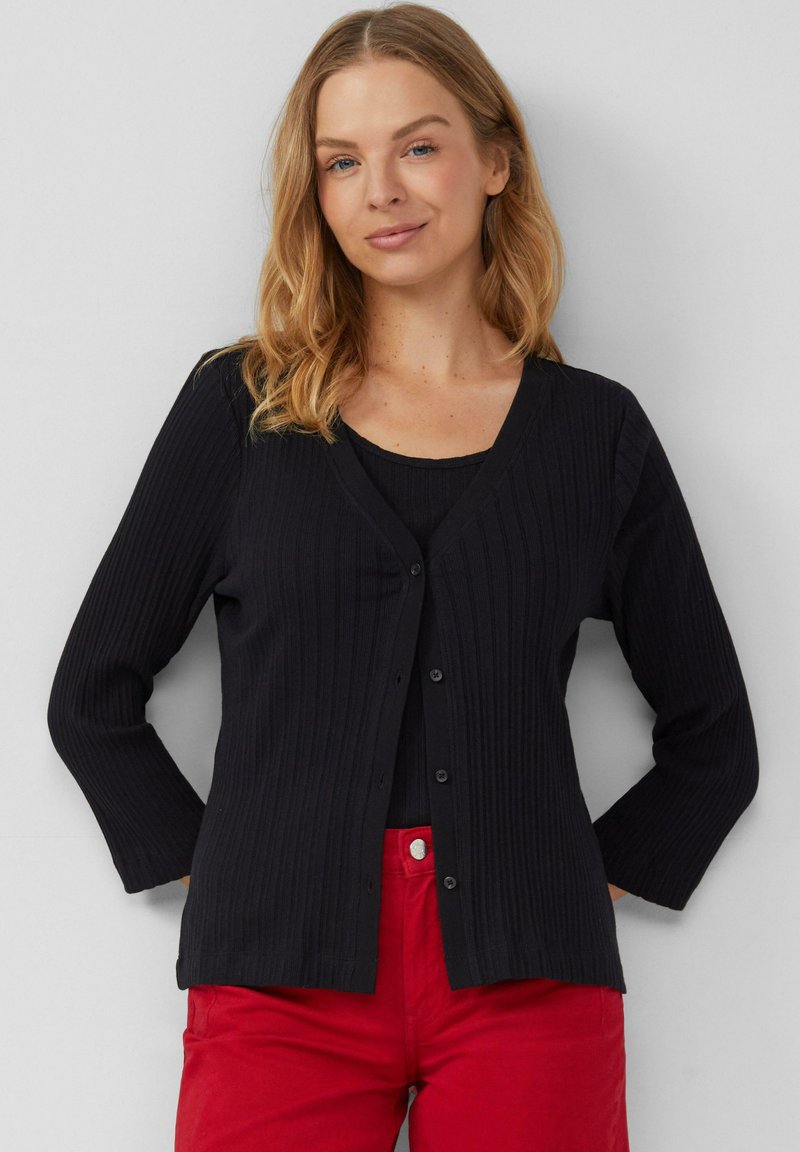 Zwarte geribde cardigan met een v-hals, lange mouwen en knopen aan de voorkant, gecombineerd met een zwarte top en rode broek. Soepele textuur en aansluitend ontwerp.