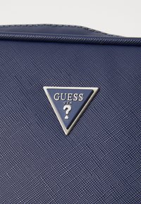 Material texturizzato blu navy con un logo metallico triangolare che riporta la scritta "GUESS" e un punto interrogativo, posizionato su uno sfondo liscio.