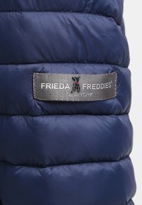 Granatowa puchowa kurtka z pikowanym teksturem. Ma srebrną metkę z napisem "FRIEDA FREDDIES" i małym logo psa na szarym tle.