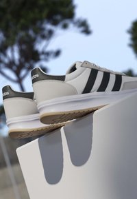 Białe i czarne sneakersy Adidas z gumowymi podeszwami, spoczywające na białej powierzchni, rozmyte tło na zewnątrz z drzewami i niebieskim niebem.
