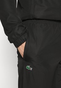 Čierne teplákové nohavice s textúrovaným povrchom, s elastickým pásom a bočnou kapsou. Pozoruhodné zelené logo krokodíla Lacoste na ľavom stehne.