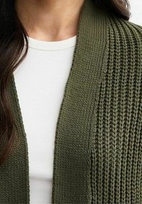 Neulejakku tummanoliivinvihreänä, jossa on teksturoitu kuvio ja avoin etuosa, kerroksittain valkoisen paidan päällä.