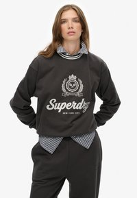Dark gray sweatshirt with a crew neckline, white logo and detailing, layered over a blue and white striped collared shirt. Casual fit.