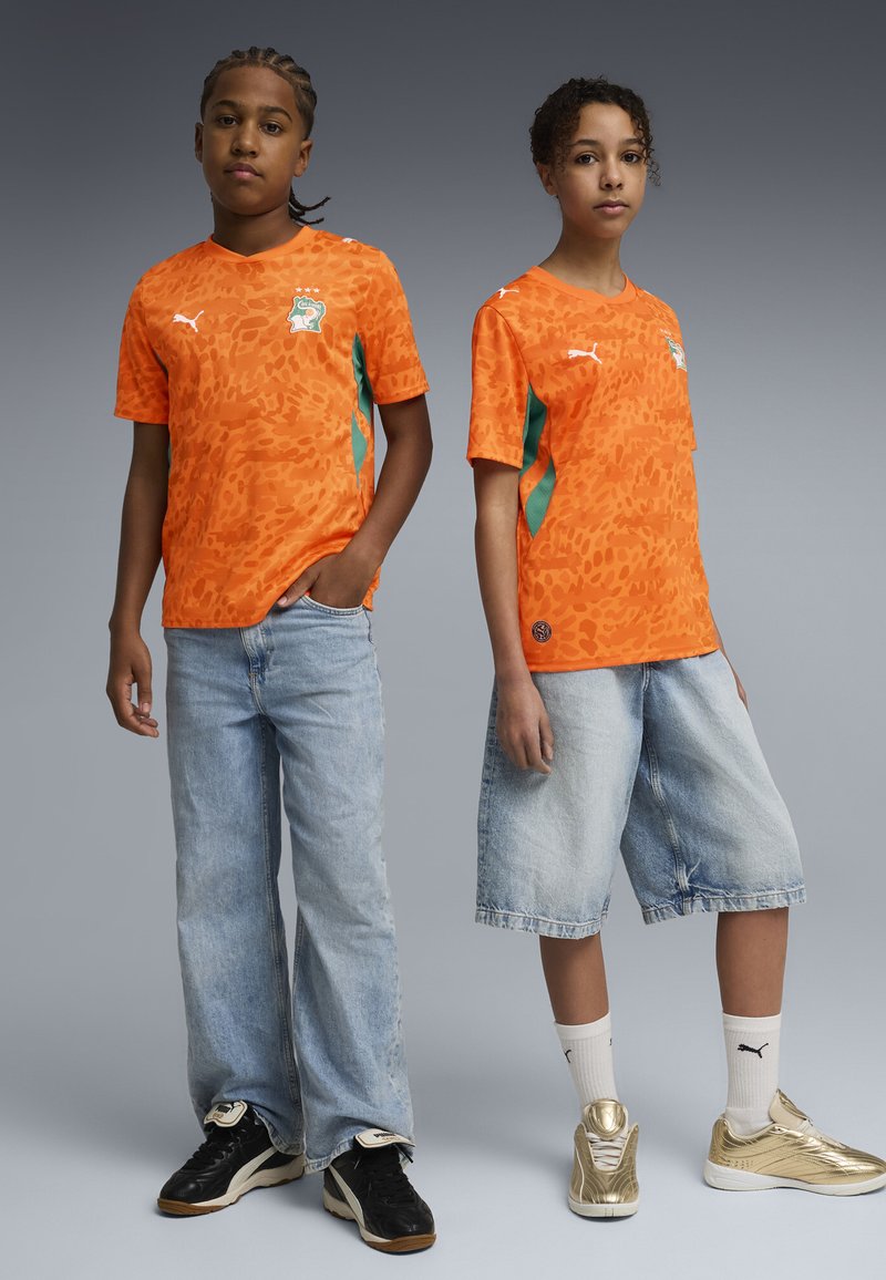 Dos jóvenes vestidos con camisetas de fútbol de patrón naranja con el logo de Costa de Marfil, pantalones vaqueros y zapatillas deportivas, posando frente a un fondo gris liso.