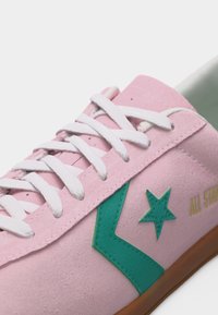 Ροζ σουέντ αθλητικό παπούτσι με λευκά κορδόνια, λογότυπο αστέρι και βέλος σε τιρκουάζ, κίτρινο κείμενο "All Star" και καφέ λαστιχένια σόλα.