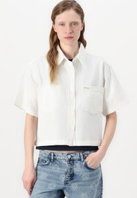 Cropped white button-up shirt made of lightweight fabric, featuring short sleeves, two chest pockets, and a subtle logo label.