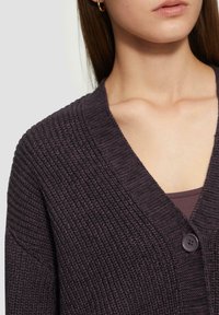 Dunkellila gestrickte Strickjacke mit V-Ausschnitt, die ein strukturiertes Muster und einen einzigen Knopfverschluss aufweist. Weiches Material, lässiges Design.
