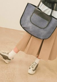Borsa a mano blu e nera con superficie testurizzata, dotata di una tasca frontale. Gonna a quadri beige abbinata a calzini bianchi e sneakers grigie e bianche.