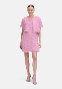 Rosa, flauschiges Kleid mit kurzen Ärmeln, knielangem Saum und strukturierten Fransen-Details. Kombiniert mit klaren High-Heels.