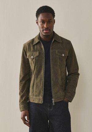 Mann trägt eine olivgrüne Wildlederjacke über einem schwarzen Hemd und dunklen Jeans, steht vor einer einfarbigen hellbeigen Wand.