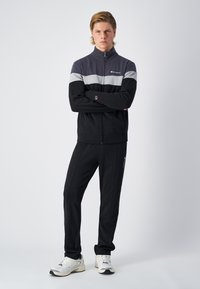 Giacca sportiva zip-up nera e grigia con colletto alto e pantaloni della tuta coordinati. Presenta il logo sul petto e strisce laterali, abbinata a sneakers bianche.