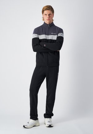 Giacca sportiva zip-up nera e grigia con colletto alto e pantaloni della tuta coordinati. Presenta il logo sul petto e strisce laterali, abbinata a sneakers bianche.