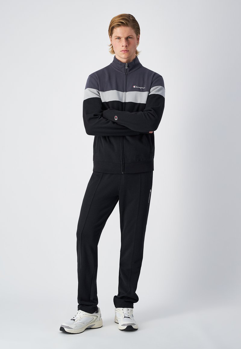 Chaqueta deportiva con cremallera negra y gris, con un cuello alto y pantalones sudadera a juego. Presenta un logo en el pecho y rayas en los lados, combinados con zapatillas blancas.