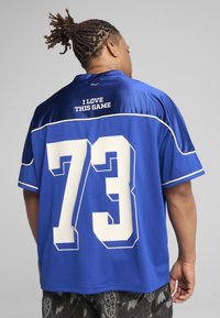 Camiseta deportiva azul con tejido de malla, detalles blancos y un gran número "73" en la parte posterior. Texto "ME ENCANTA ESTE JUEGO" por encima del número.