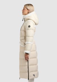 Beige gewatteerde jas met een pluizige witte kraag en manchetten, voorzien van een riem, zijrits en logodetail op de mouw. Halflang ontwerp.
