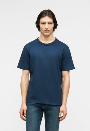 Młody mężczyzna o średniej długości ciemnych włosach, ubrany w prostą granatową koszulkę z krótkim rękawem i niebieskie dżinsy, stojący na tle białego tła.