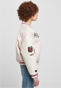 Lys rosa bomberjakke med hvite ermer, ribbede mansjetter og røde detaljer. Har FUBU-merke og broderte detaljer. Teksturert stoff.