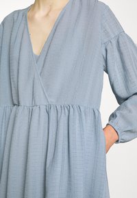 Femme portant une robe bleu clair texturée à manches longues avec un col en V et une taille froncée, main placée dans la poche latérale, sur un fond uni.