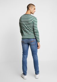 Camisa de manga larga a rayas verdes y grises, estilo ajustado. Lleva puesta unos vaqueros en azul claro. La camisa tiene un escote redondo y un corte slim.